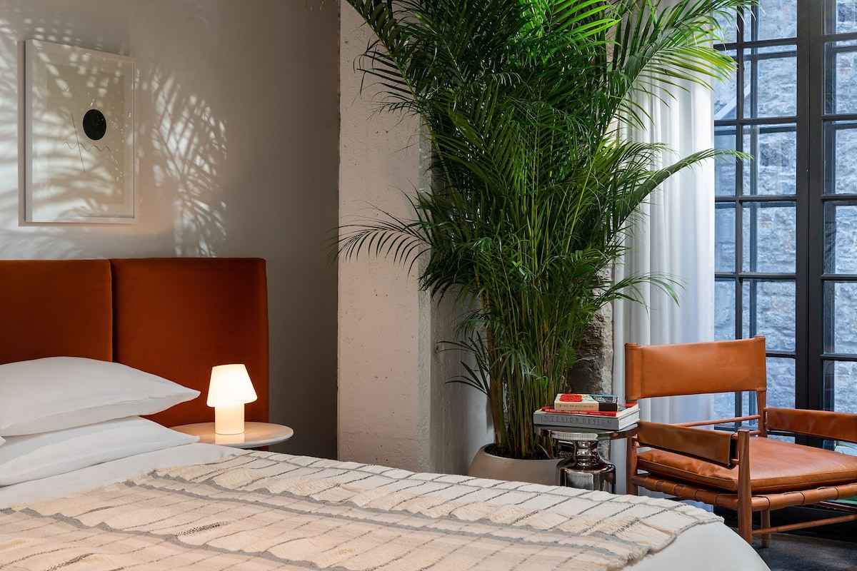 Una cama con ropa de cama blanca, una lámpara de noche, una planta grande en maceta, un sillón de cuero y una ventana con vistas a árboles.