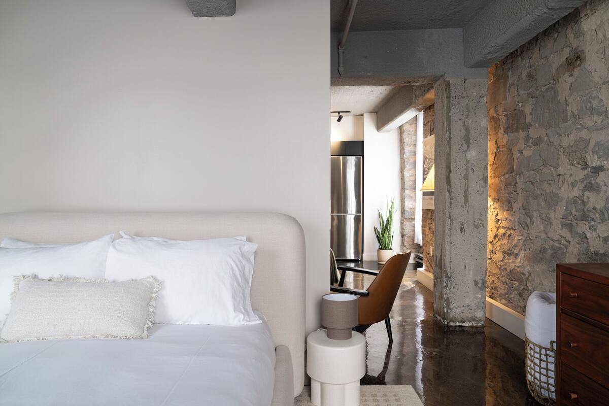 A bedroom with a bed, a chair, a dresser, and a view of a kitchen area.
