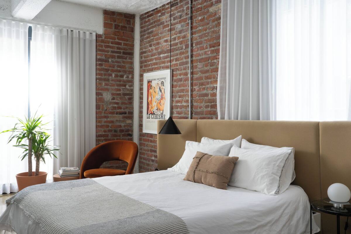A hotel room with a brick wall, a bed with white bedding, a brown headboard, a potted plant, and a framed poster on the wall.
