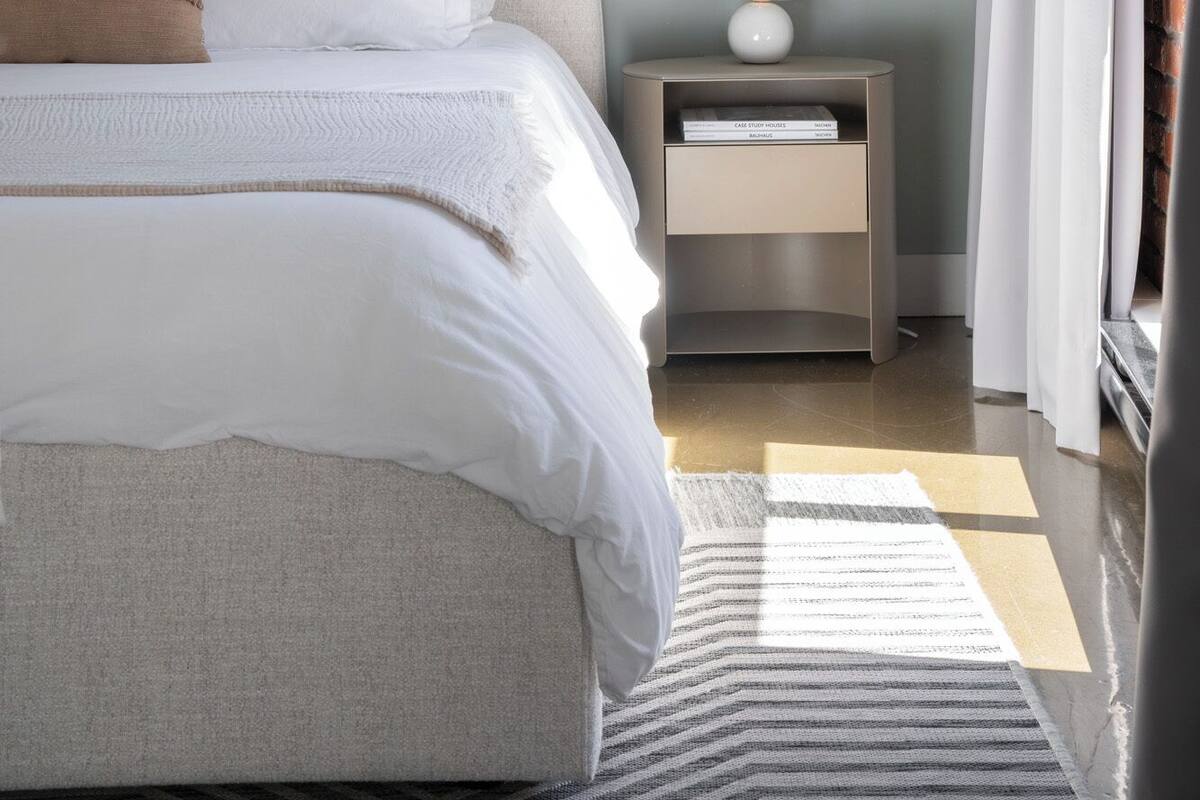 A neatly made bed with white linens and a beige throw blanket, a bedside table with a lamp and a book, and a striped rug on the floor.