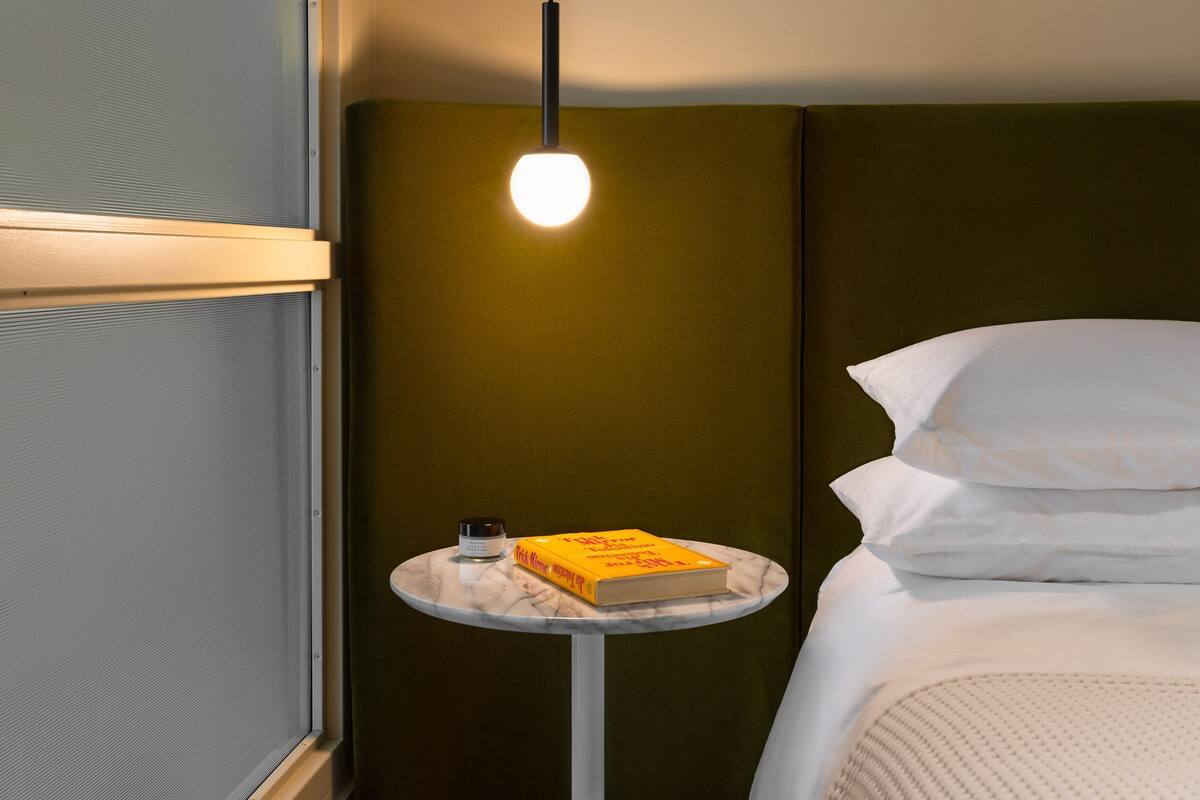A hotel room with a bed, a round table with a book, a hanging light, and a window with blinds.