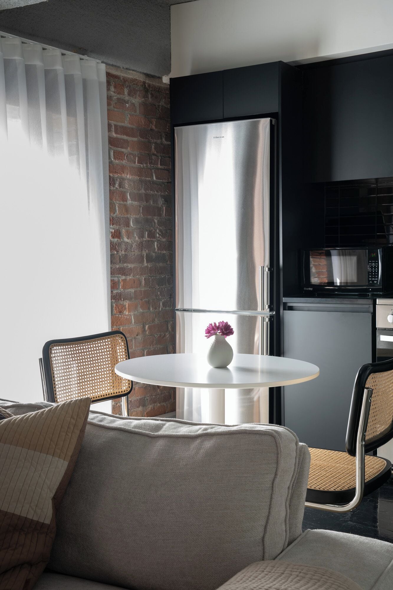 A modern kitchen with a stainless steel refrigerator, a microwave, and a small oven. There's a round white table with a vase of flowers. The room has a brick wall and a window with sheer curtains.