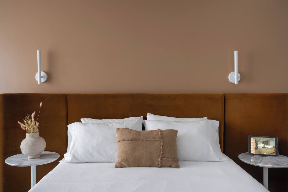 A neatly made bed with white linens and a brown accent pillow, flanked by two small round tables with a vase of dried flowers on one and a tablet on the other.