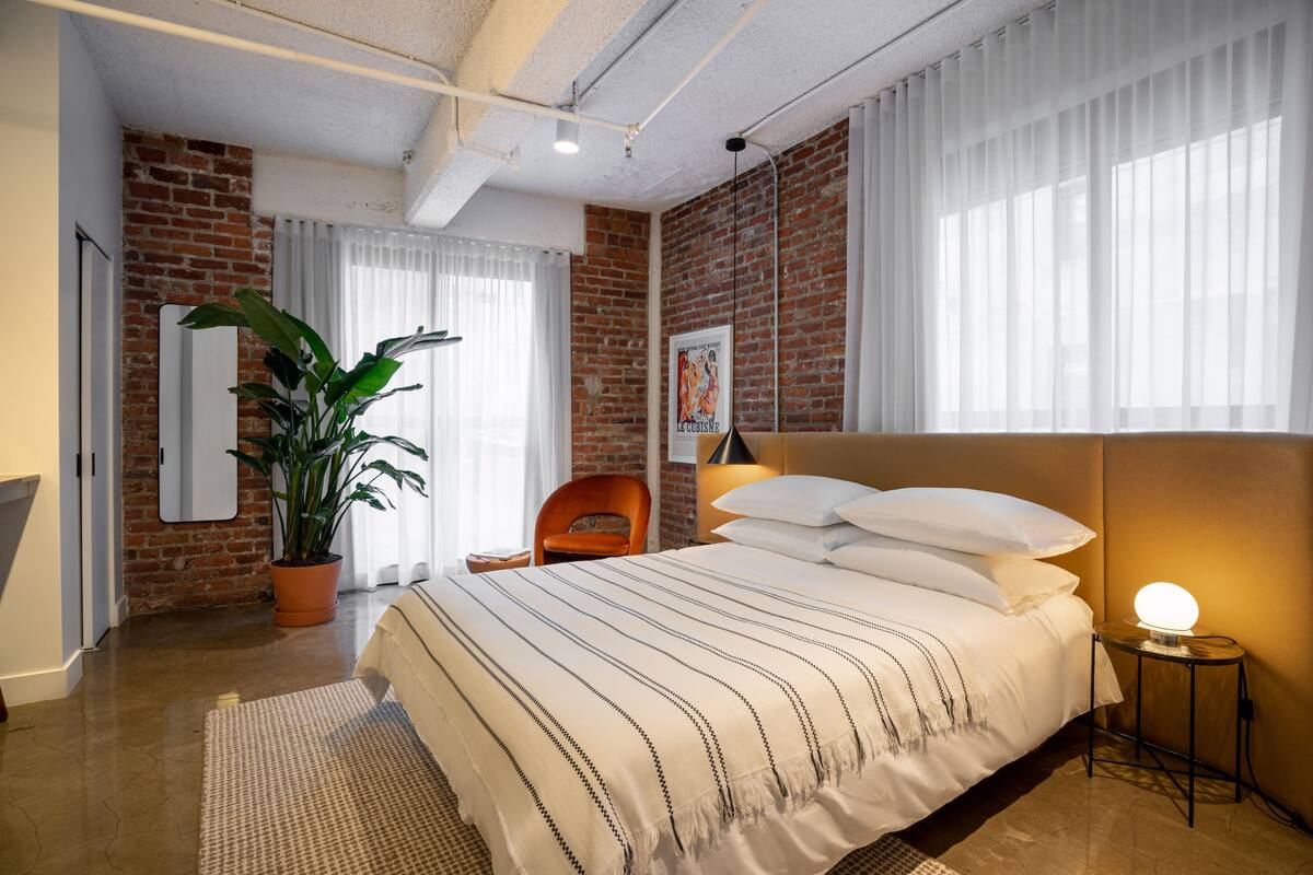 A hotel room with a brick wall, a bed with striped bedding, a potted plant, a chair, and a bedside lamp.