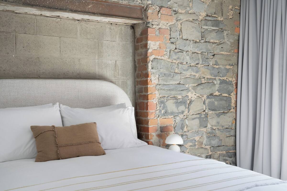 A bed with white bedding and a brown pillow, a stone wall, and a lamp.