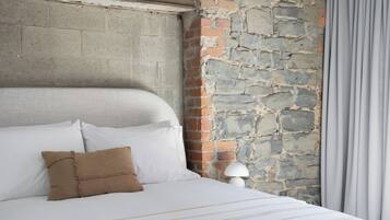 Una cama con ropa de cama blanca y un almohadón marrón, una pared de piedra y una lámpara.
