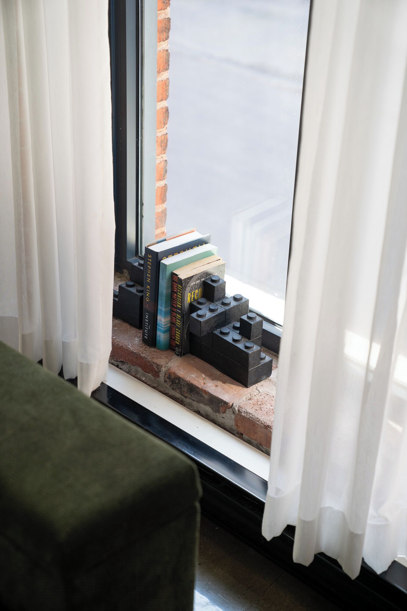 A window with white curtains, a brick wall, and a small step with books and LEGO bricks.