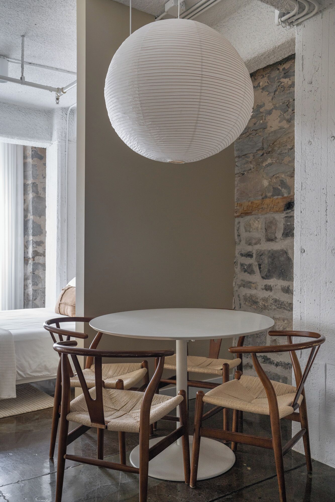 Una mesa redonda para comer con cuatro sillas de madera, una lámpara colgante blanca y una pared de piedra al fondo.