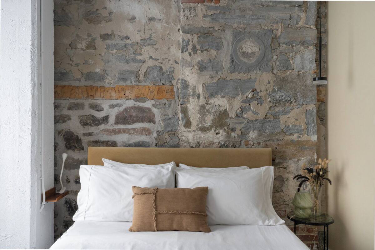 A bedroom with a stone wall, a wooden headboard, white bedding, and a small bedside table with a vase.