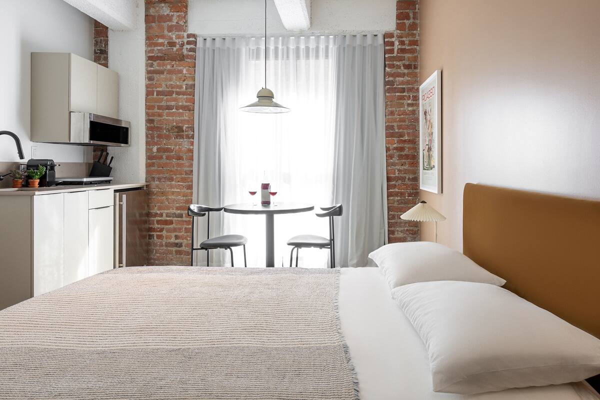 A hotel room with a bed, a dining table with chairs, a kitchen area with a microwave, and a brick wall.