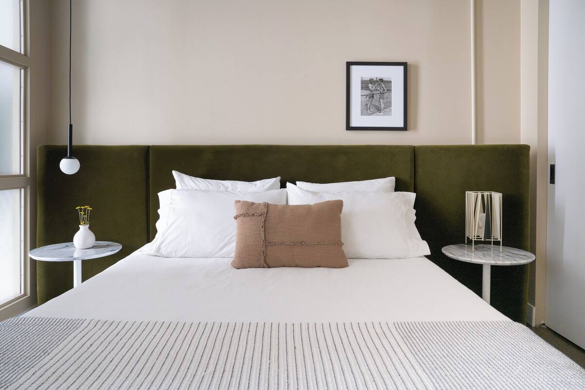 A neatly made bed with white linens and a brown accent pillow, a framed picture on the wall, and a small round table with a vase of flowers.