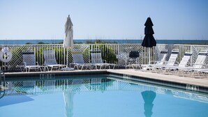 A pool with white lounge chairs and umbrellas, a clear view of the ocean, and a lifeguard tower.