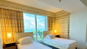 Ein Hotelzimmer mit zwei Betten, großen Fenstern mit Vorhängen, Holzboden und Meerblick.