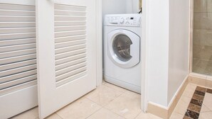 Ein kleiner Hauswirtschaftsraum mit einer Waschmaschine mit Frontlader, einer Tür mit verstellbaren Lamellen und einem Fliesenboden.