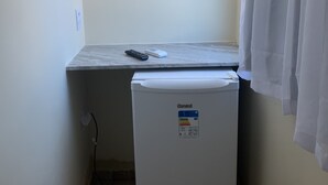 A small, white refrigerator with a control panel, a remote control, and a white object on top of it, placed in a corner of a room with a wooden floor and a white curtain.