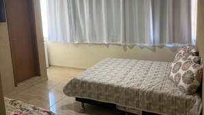 A bedroom with a bed, a window with white curtains, a door, and a tiled floor.
