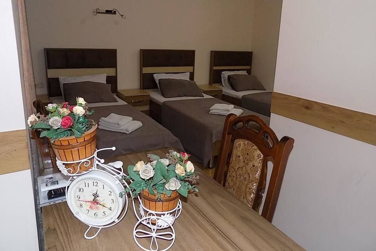 Ein Zimmer mit drei Betten, einem Holztisch mit einer Uhr und Blumenkörben sowie einem Stuhl.