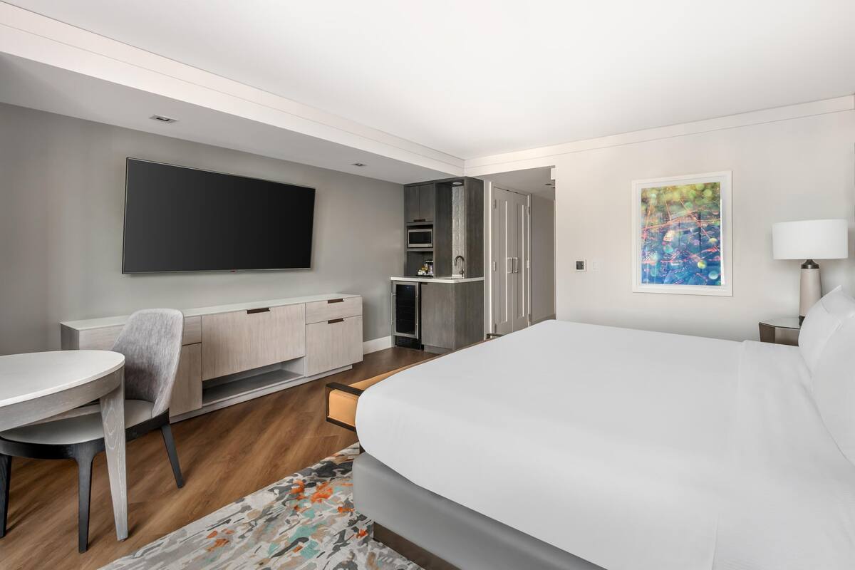 Una habitación de hotel moderna con televisión de pantalla plana, una cama, un escritorio con una silla, un microondas y un armario.
