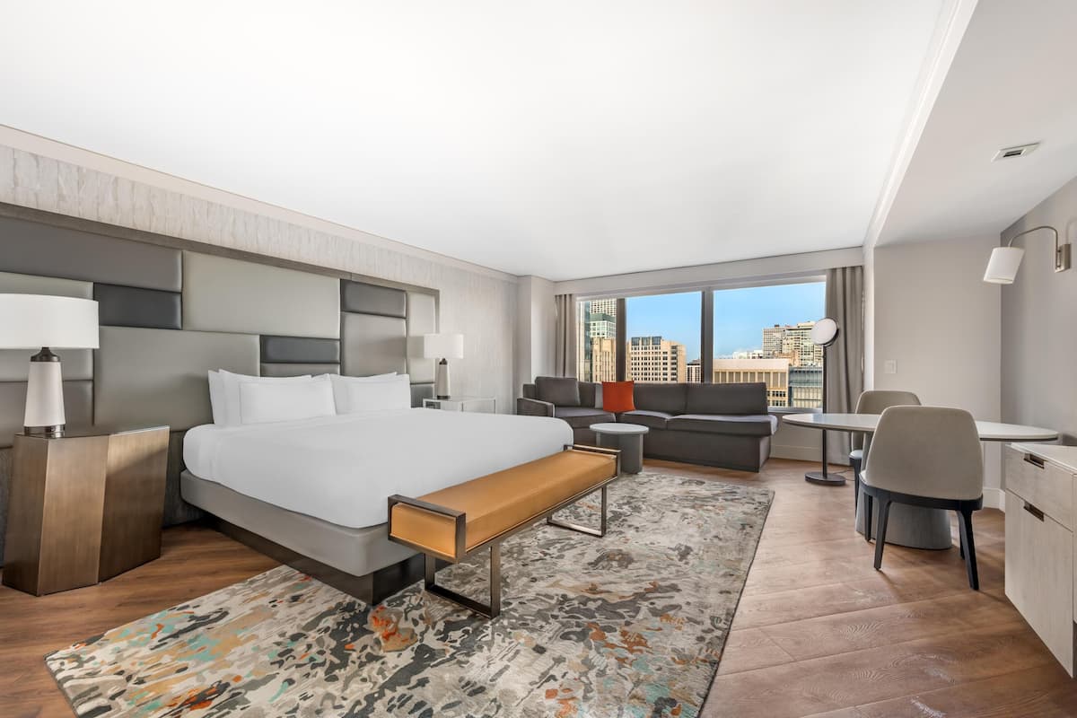 Una habitación de hotel moderna con una cama grande, un sofá, una mesa de comedor y un escritorio. Se ve la ciudad a través de amplias ventanas.