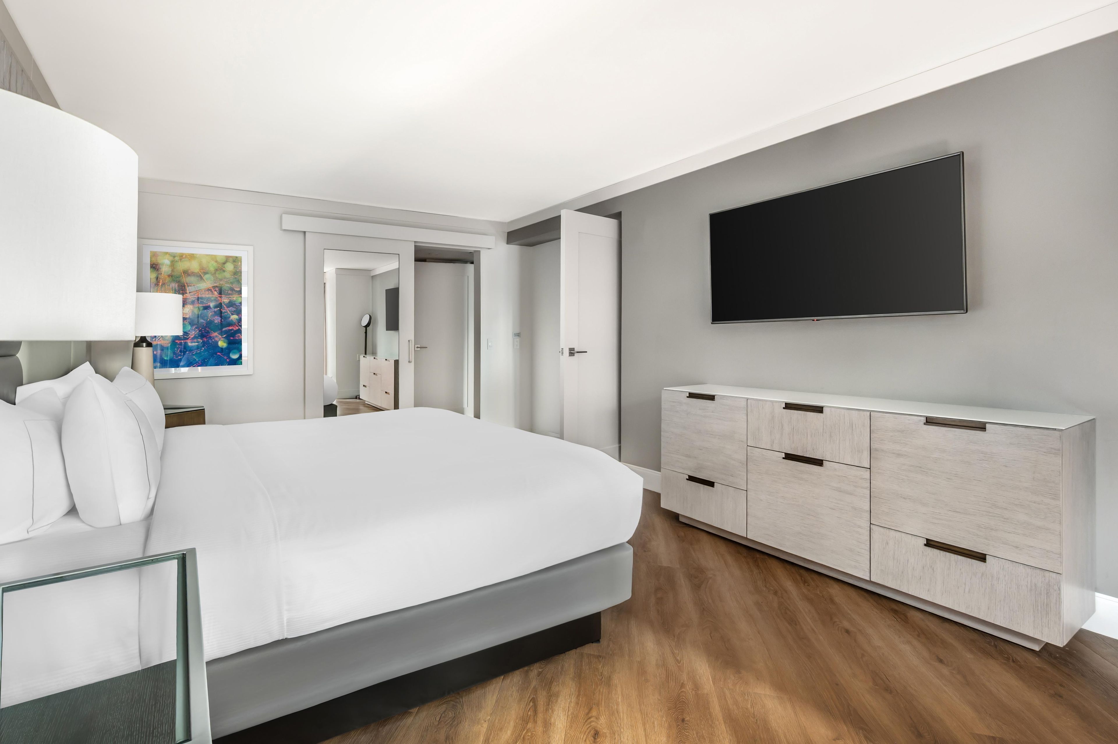 A modern bedroom with a large bed, a flat-screen TV mounted on the wall, and a minimalist dresser.
