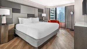 Una habitación de hotel moderna con una cama grande, dos mesitas de noche con lámparas, una pequeña mesa con una silla, un ventanal con vistas a la ciudad y un televisor de pantalla plana.