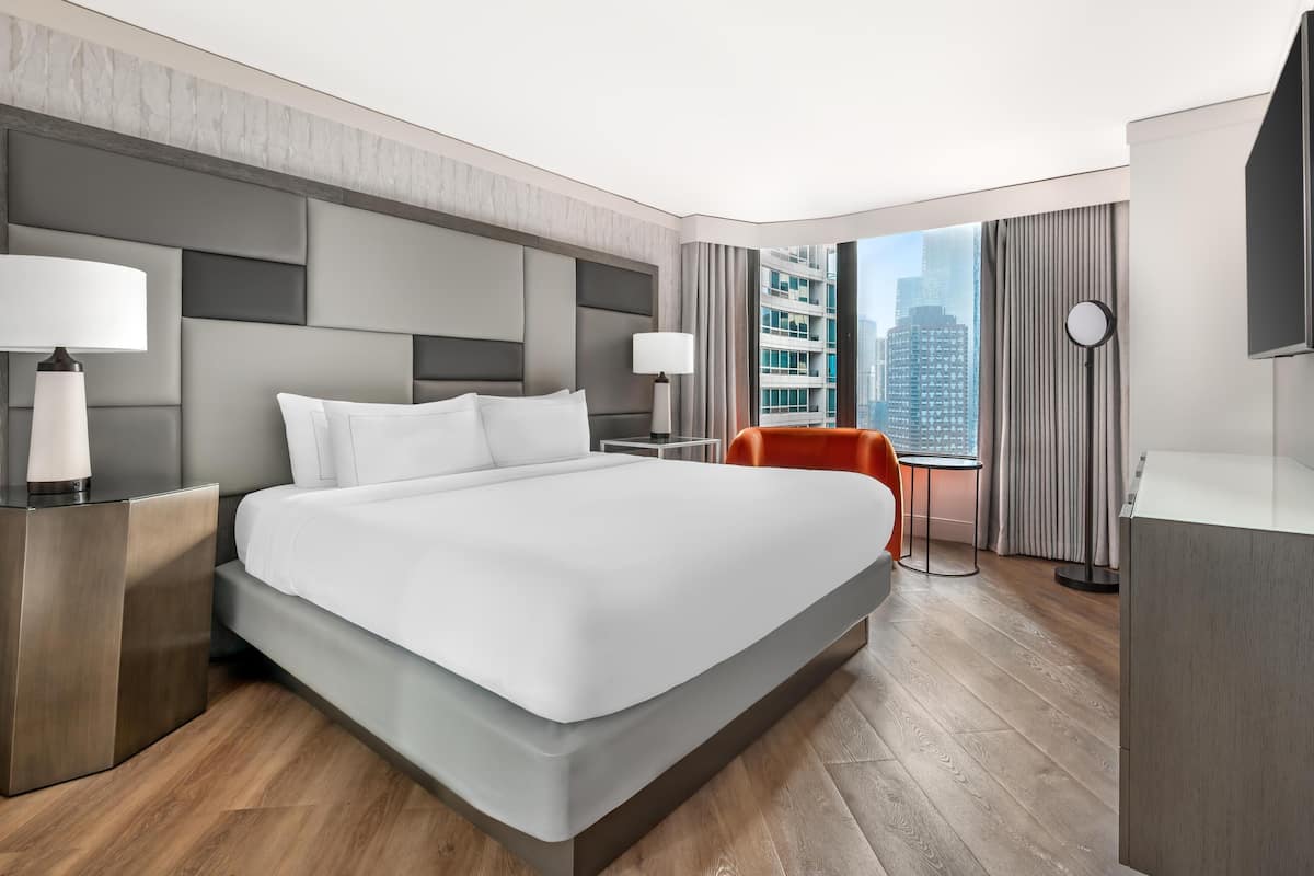 Una habitación de hotel moderna con una cama grande, dos mesitas de noche con lámparas, una pequeña mesa con una silla, un ventanal con vistas a la ciudad y un televisor de pantalla plana.