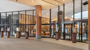Un moderno vestíbulo de hotel con amplias puertas de cristal, columnas de madera y plantas en macetas.