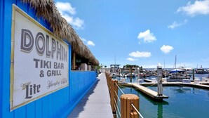 A dockside view of Dolphin Tiki Bar & Grill with a thatched roof, a wooden walkway, and a marina in the background.