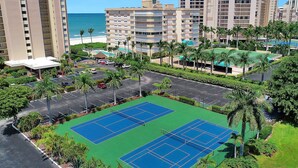 Un court de tennis entouré de palmiers et un parking, avec une plage et des immeubles de grande hauteur en arrière-plan.