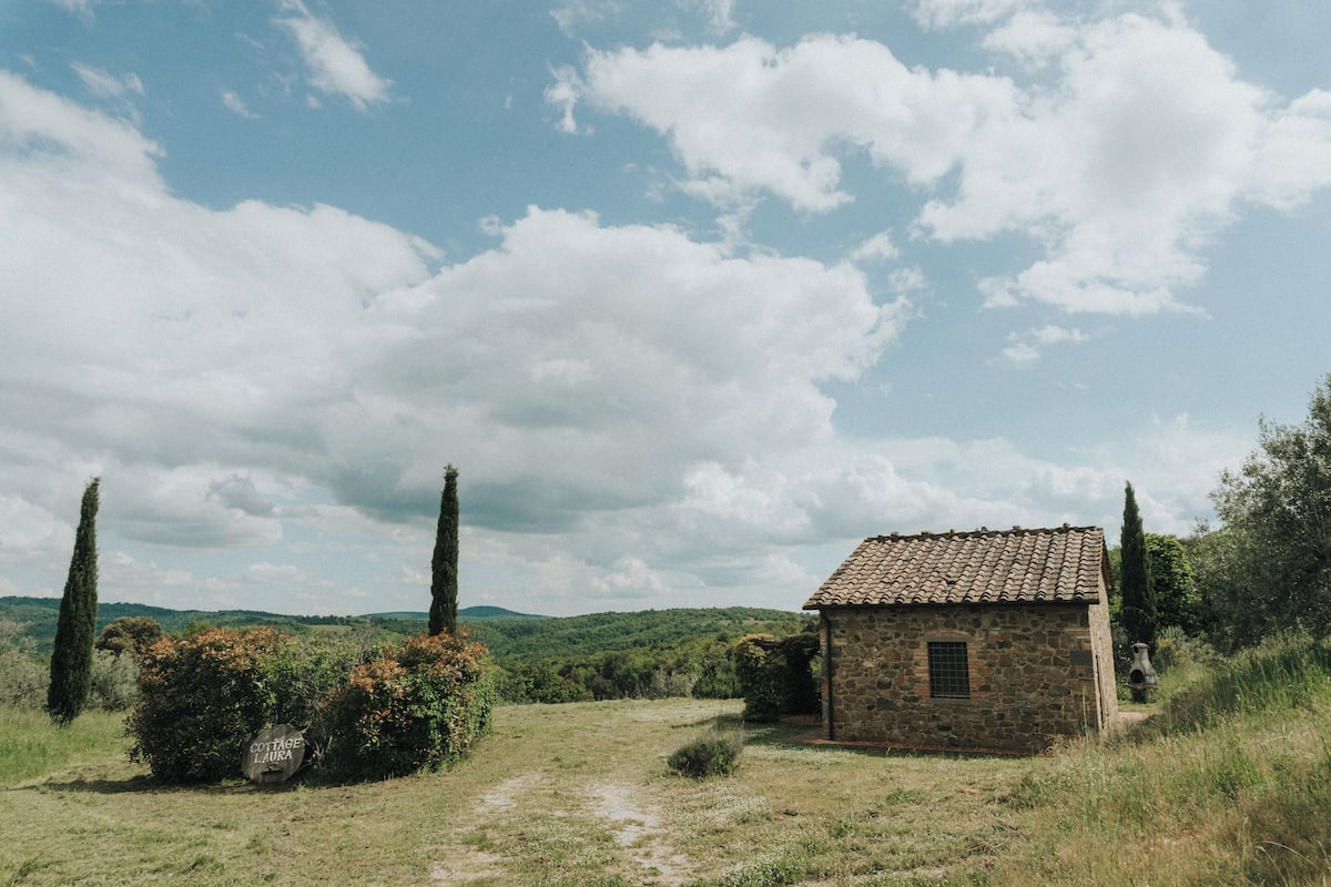 A rustic stone cottage with a tiled roof, surrounded by lush greenery and a clear sky.