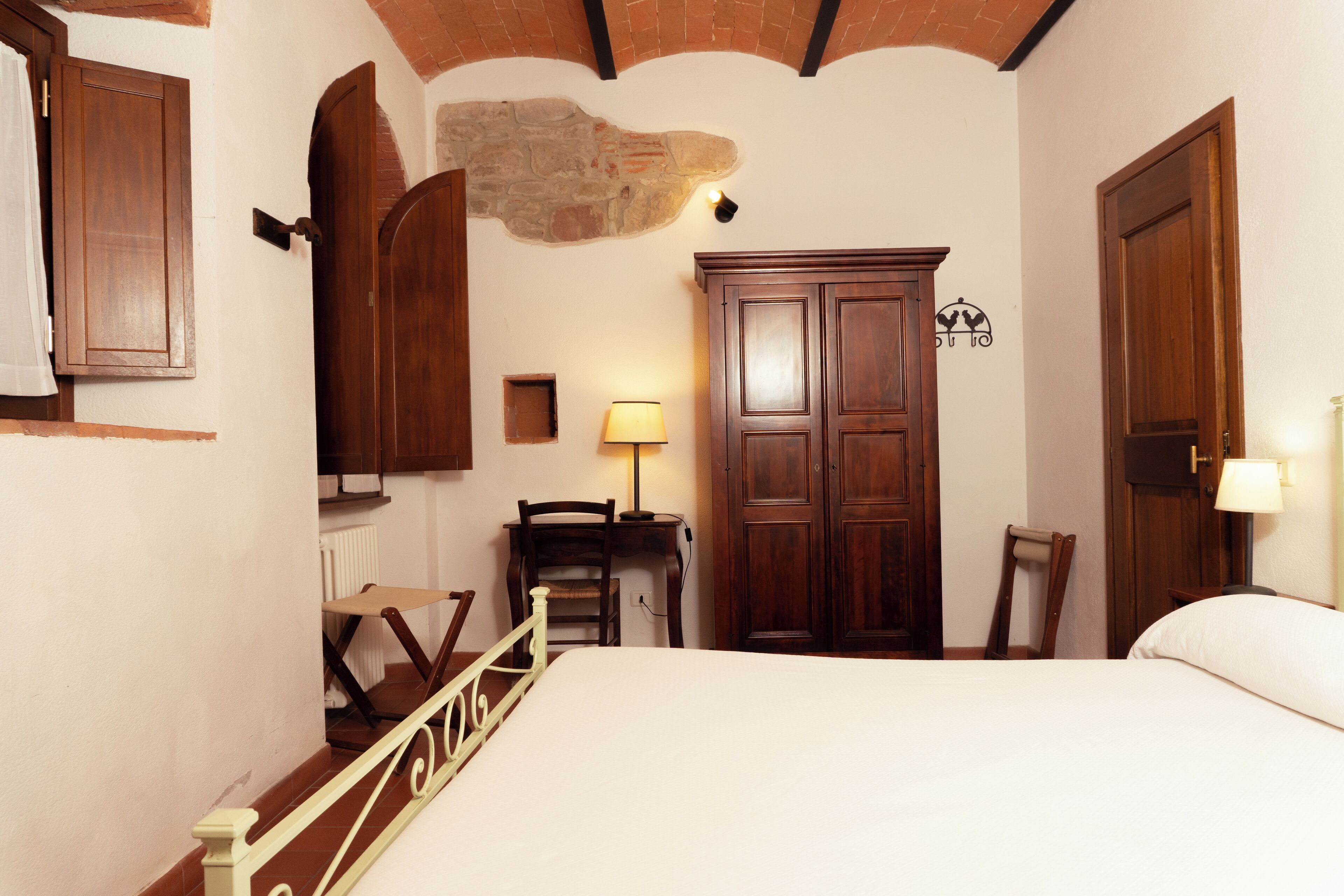 A bedroom with a bed, wooden wardrobe, desk, chair, and a stone wall.