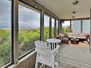 A covered outdoor area with a view of the beach, wooden deck, white chairs, a round table, and a sofa. - Sea Strapped | Oceanfront + Swim Spa (Topsail Beach)