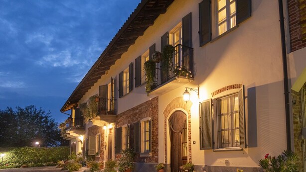 Un edificio de dos plantas con fachada de piedra, múltiples ventanas con contraventanas y un balcón con plantas.