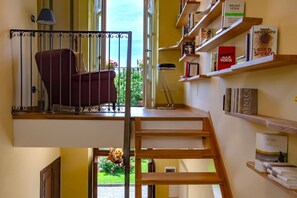 Una habitación con una silla roja, una lámpara, una estantería llena de libros y vistas al exterior con zonas verdes.