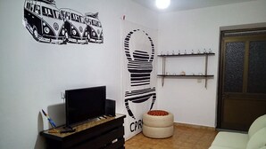 Un salon avec un téléviseur posé sur un meuble, un canapé, une petite table avec des magazines, un élément décoratif mural et un tabouret rond.