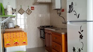 Une cuisine compacte équipée d’un four à micro-ondes, d’une cuisinière et d’un réfrigérateur. On y trouve une poubelle verte et un meuble en bois avec des tiroirs.