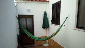 Un couloir étroit avec un hamac vert, une porte ornée d’un panneau de verre décoratif et une fenêtre avec des volets marron.