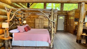 A log cabin bedroom with a bed under a wooden archway, wooden floors, and a door leading to another room. - Elkmont Views - 1BR/New Hot Tub/New Firepit/Grill/Sleeps 6 (Gatlinburg)