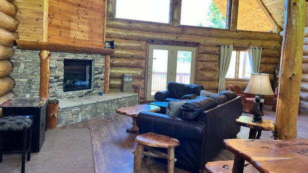 A log cabin interior with a stone fireplace, wooden furniture, and large windows. - Elkmont Views - 1BR/New Hot Tub/New Firepit/Grill/Sleeps 6 (Gatlinburg)