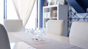 Un coin repas avec un grand saladier en verre transparent posé sur une table, des chaises blanches, une nappe blanche, une bibliothèque et un mur bleu orné d’un motif géométrique.