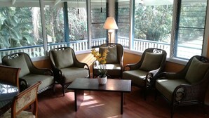 A sunlit room with wicker furniture, a central table with a potted plant, and large windows offering a view of greenery outside.