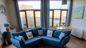 Ein Wohnzimmer mit blauem Ecksofa, einem grauen Teppich, Holzböden, einem Esstisch mit Stühlen und großen Fenstern mit Vorhängen.