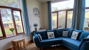 Ein Wohnzimmer mit einem blauen Modulsofa, einem hellblauen Teppich, Holzdielen und großen Fenstern mit Vorhängen.