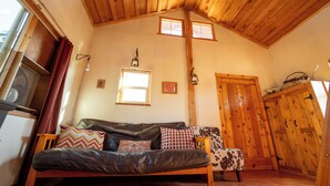 A cozy room with a wooden ceiling, a leather sofa with patterned cushions, a small wooden chair, and a wooden door with a basket on top. - Glamping, off-grid adventure at peace-friendly cabin on 10 acres w/ STARGAZING! (Fredonia)