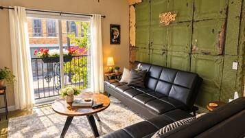 A living room with a black leather sofa, a round coffee table with books and a remote, a balcony with plants, and a green wall with a 'Tiger' sign.