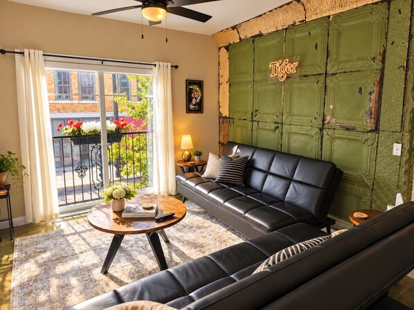 Ein Wohnzimmer mit einer schwarzen Ledersofa, einem runden Couchtisch mit Büchern und einer Fernbedienung, einem Balkon mit Pflanzen und einer grünen Wand mit einem 'Tiger'-Schild.
