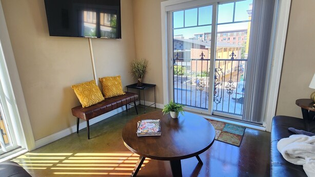 A modern living room with a sofa, a round table, a TV mounted on the wall, and a balcony with a view of buildings. - 9th St. Beals Loft Downtown Columbia (Columbia)