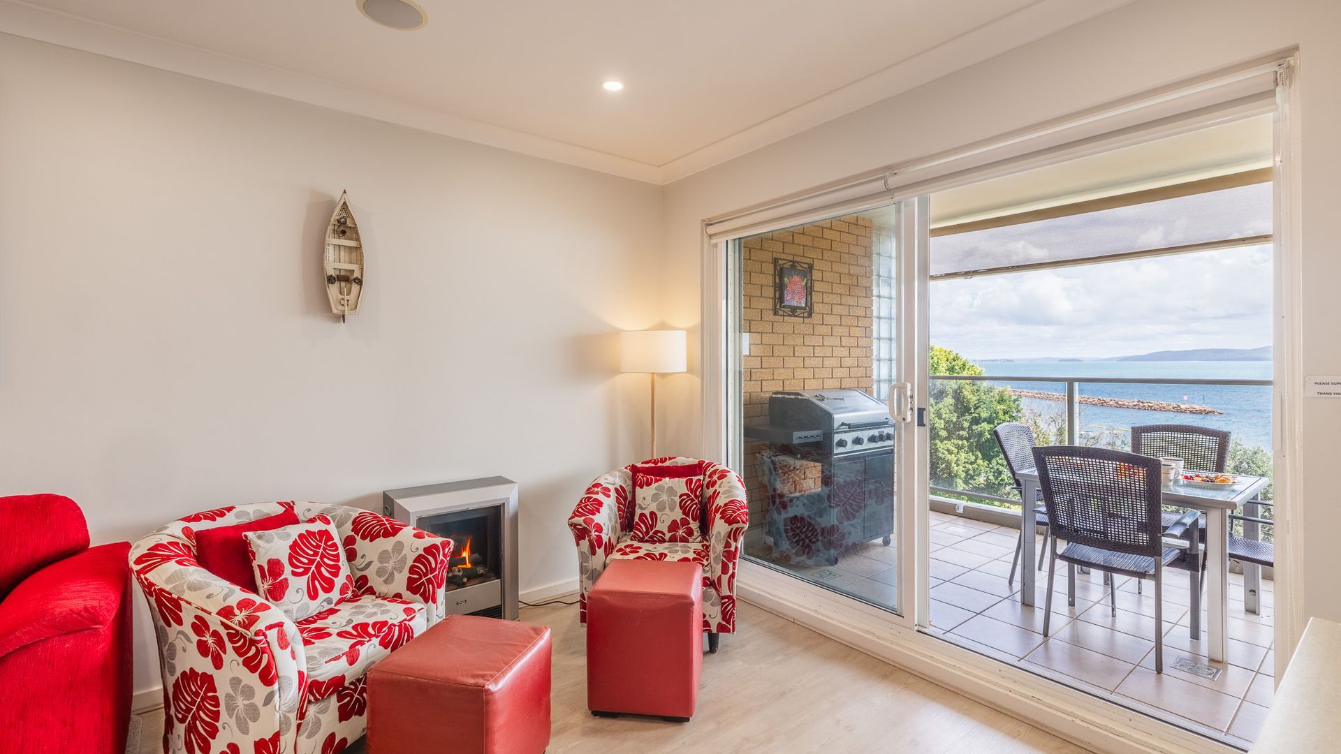 A modern living room with a fireplace, a sofa, a chair, and a table. There is a balcony with a grill and a view of the sea.