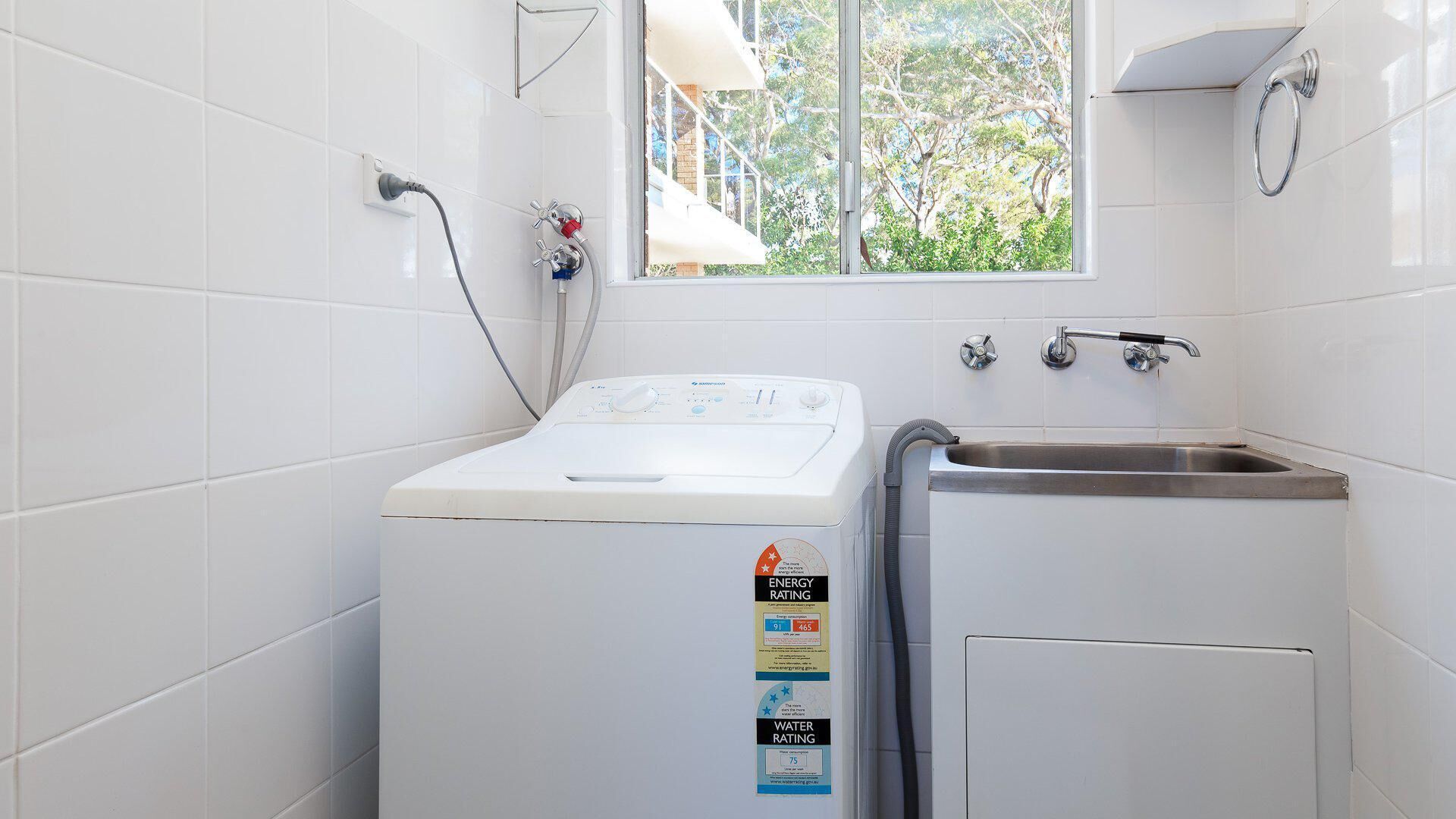 Een wasruimte met een voorlader wasmachine, een wastafel en een raam met uitzicht op bomen.