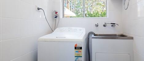 Een wasruimte met een voorlader wasmachine, een wastafel en een raam met uitzicht op bomen.